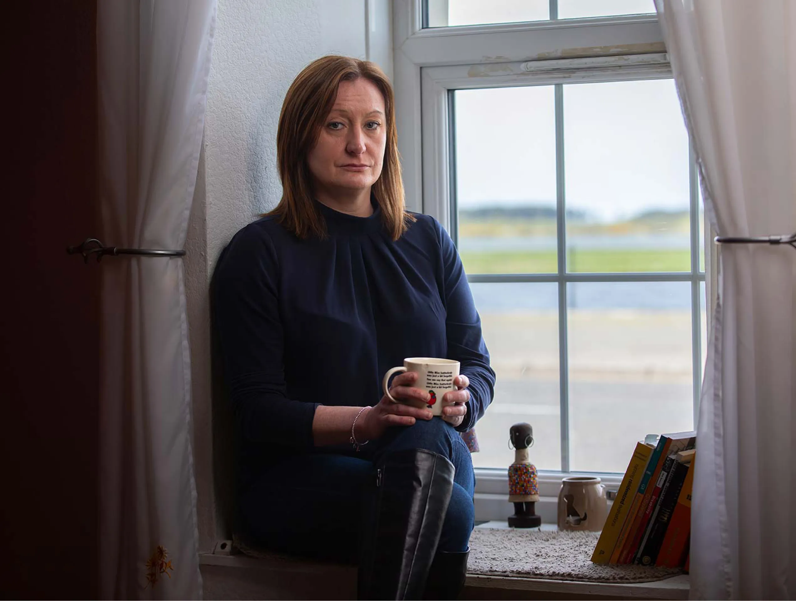 Portrait photo of a female UK Armed Forces veteran sitting on a windowsill holding a mug.