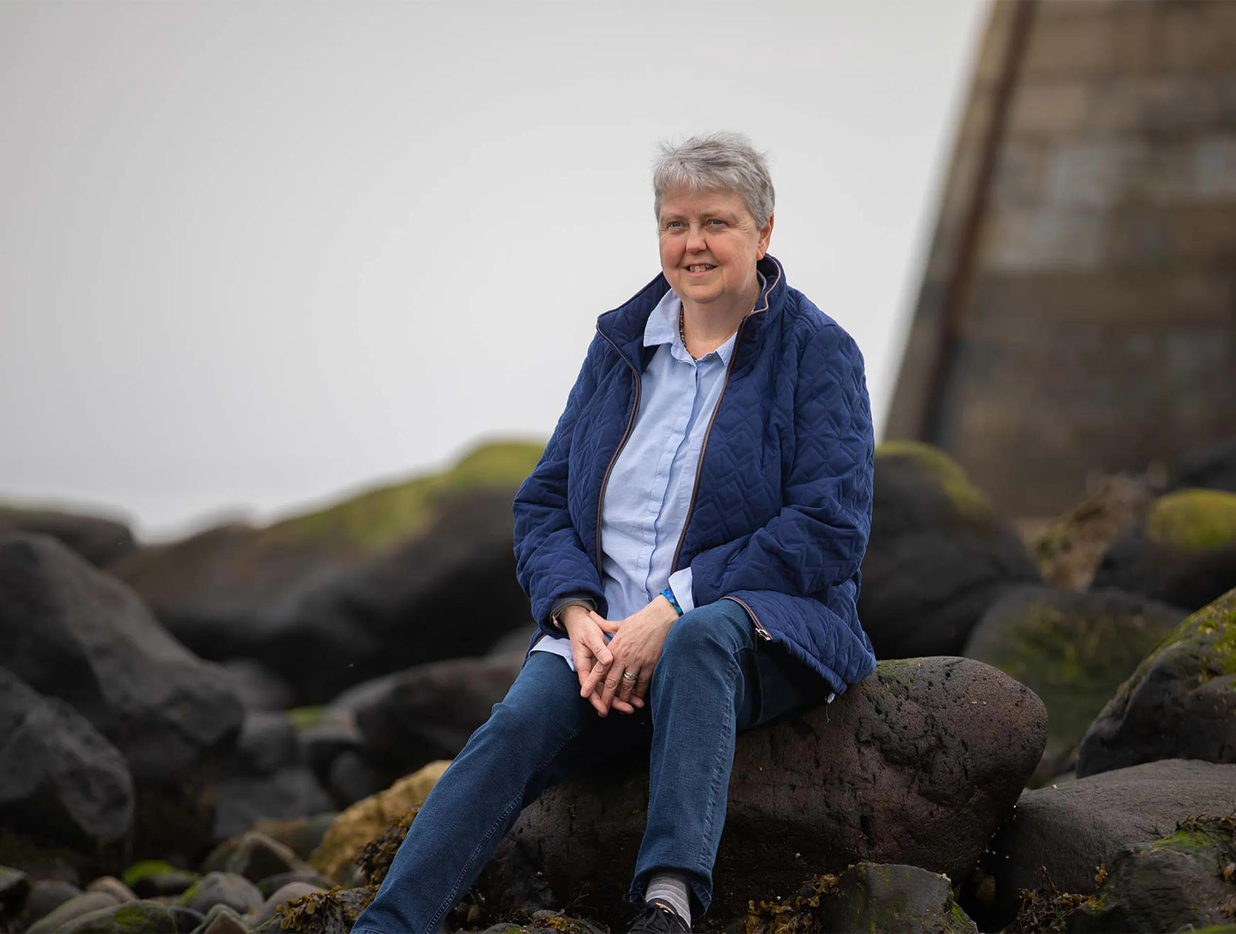 Portrait photo of a female UK Armed Forces veteran sitting on a rock near the coast.