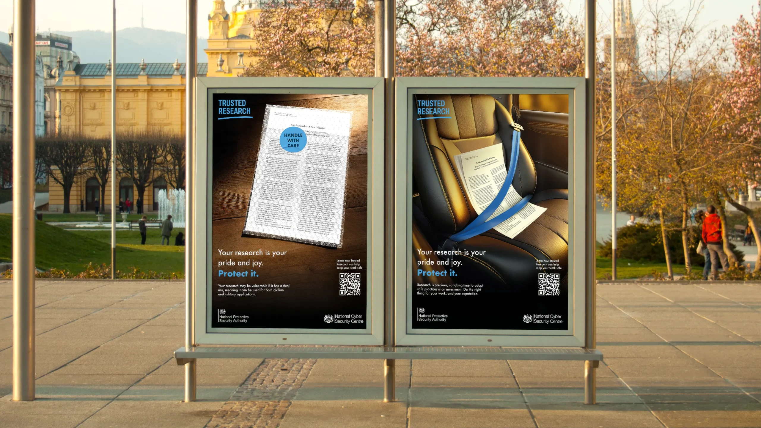 A bus shelter displays two Trusted Research campaign posters in an outdoor public space. One poster shows a research paper wrapped in protective bubble wrap with a "Handle with Care" sticker, while the other shows a research paper secured with a seatbelt in a car seat. Both posters feature the message, "Your research is your pride and joy. Protect it." alongside the Trusted Research logo, a QR code, and National Protective Security Authority and National Cyber Security Centre branding. Trees, historic buildings and pedestrians are visible in the background.