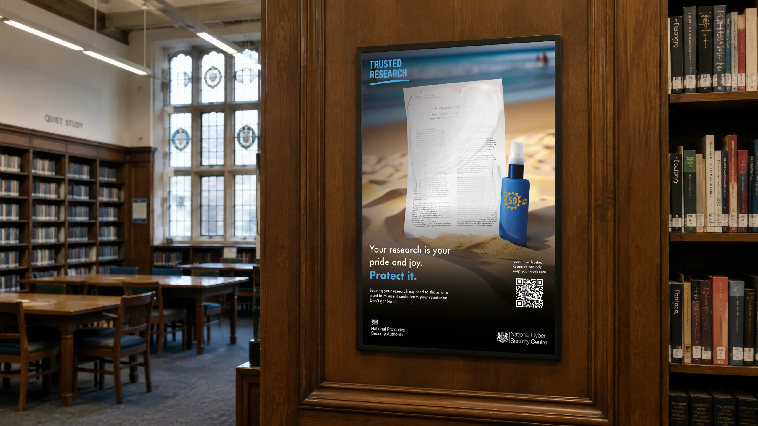 A framed Trusted Research campaign poster is displayed on a wooden panel inside a university library, with bookshelves, study tables and large windows in the background. The poster shows a research paper standing on a sandy beach with sunscreen being applied from a bottle of sun cream, reinforcing the message, "Your research is your pride and joy. Protect it." It includes the Trusted Research logo, a QR code, and National Protective Security Authority and National Cyber Security Centre branding.